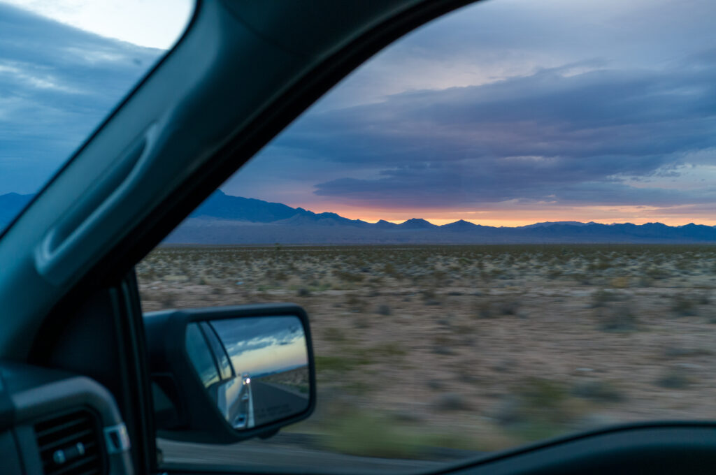 Blick aus einem Fahrzeugfenster auf eine weite Wüstenlandschaft mit Bergen im Hintergrund bei Sonnenaufgang.